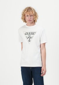 Valkoinen puuvillainen t-paita, jossa on suuri kolmion muotoinen "GUESS"-logo mustana ja hopeisena, yksityiskohtaisella suunnittelulla, yhdistetty tummansinisiin farkkuihin.