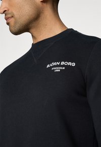 Mann trägt schwarzes Sweatshirt mit Rundhalsausschnitt und weißem "Björn Borg Stockholm 1956"-Logo auf der Brust, dargestellt von der Schulter bis zum Kinn.