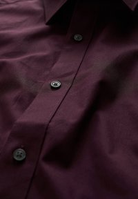 Burgundy knapp-up skjorta med en slät textur, med en spetsig krage och svarta knappar längs knappslaget.