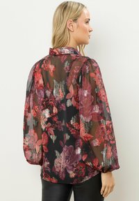Transparente schwarze Bluse mit roten und lila Blumenmustern, gepufften Ärmeln und gerafften Bündchen. Glatter, leichter Stoff mit einem leichten Glanz.