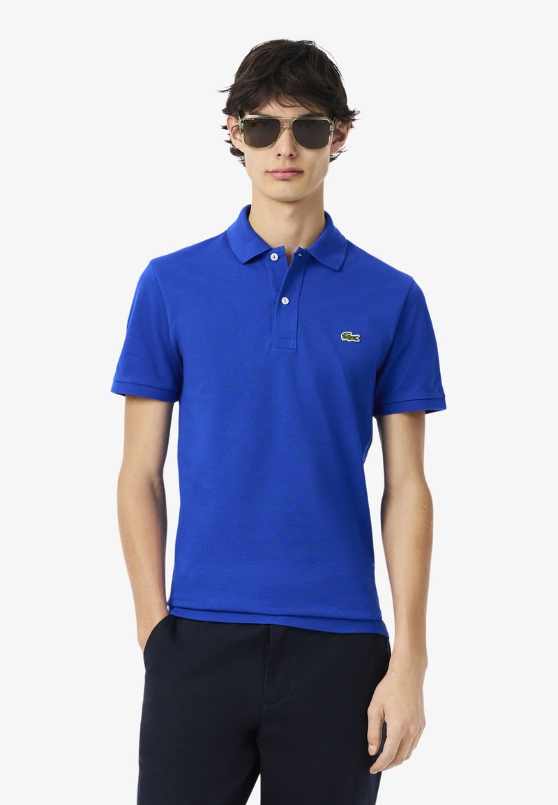 Blå polo skjorta i bomull, med klassisk krage, treknappslås och en liten grön krokodil-logotyp på vänster bröst.