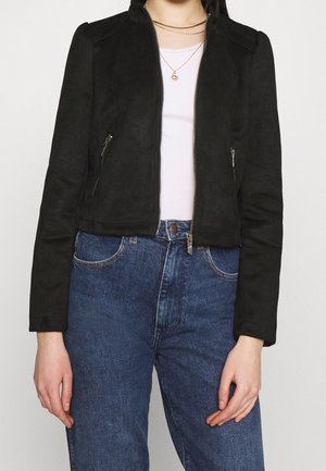 Person trägt eine schwarze Reißverschlussjacke über einem weißen Hemd, blaue hochgeschnittene Jeans und geschichtete goldene Halsketten.