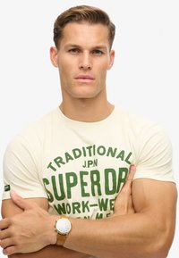 Camiseta crema con texto gráfico verde; material de algodón suave; mangas cortas; llevada con un reloj marrón en una muñeca musculosa.