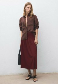 Hnědá košile na knoflíky, burgundská maxi sukně, černá taška přes rameno a černé sandály. Jednoduchý design, hladká tkanina a uvolněný střih.