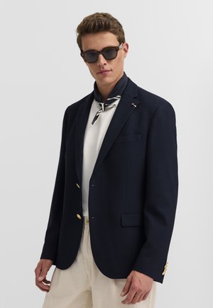 Mann trägt dunkelblauen Blazer, weißes Hemd, beige Hose, gestreiften Halstuch und Sonnenbrille, steht vor schlichtem hellem Hintergrund.