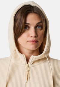 Felpa con cappuccio a coste di colore crema, con zip integrale, lacci regolabili e texture morbida. Presenta un ampio cappuccio e cuciture dettagliate.