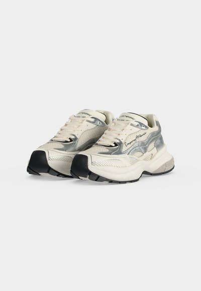 Ζευγάρι λευκά και ασημί chunky sneakers με μαύρες σόλες και ανοιχτό μπεζ κορδόνια σε απλό ανοιχτό φόντο.