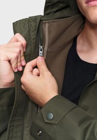 Zelená nepremokavá bunda so zipsom vpredu, vrstvenou fleecovou podšívkou a zapínaním na gombíky na manžetách. Ruky držia zips na zapnutie.