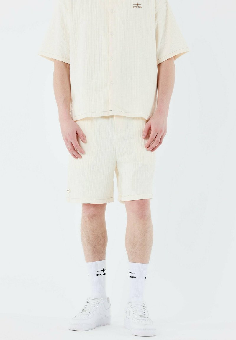 Camiseta de manga corta color crema y pantalones cortos a juego, tejido texturizado con rayas verticales; combinados con zapatillas blancas y calcetines negros con logotipo.