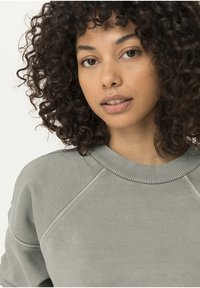 Sudadera gris con un cuello redondo acanalado y mangas raglán. Hecha de una tela suave con una textura lisa. Cabello rizado natural visible.