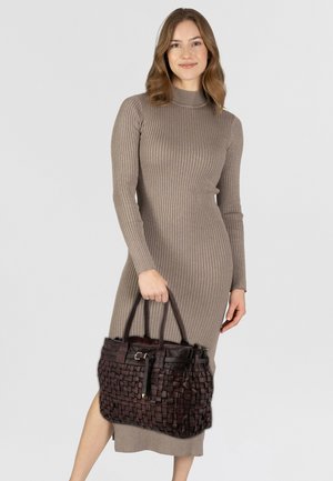 Una donna che indossa un vestito taupe a maniche lunghe a coste tiene una grande borsa a tracolla in pelle intrecciata di colore marrone scuro su uno sfondo chiaro.