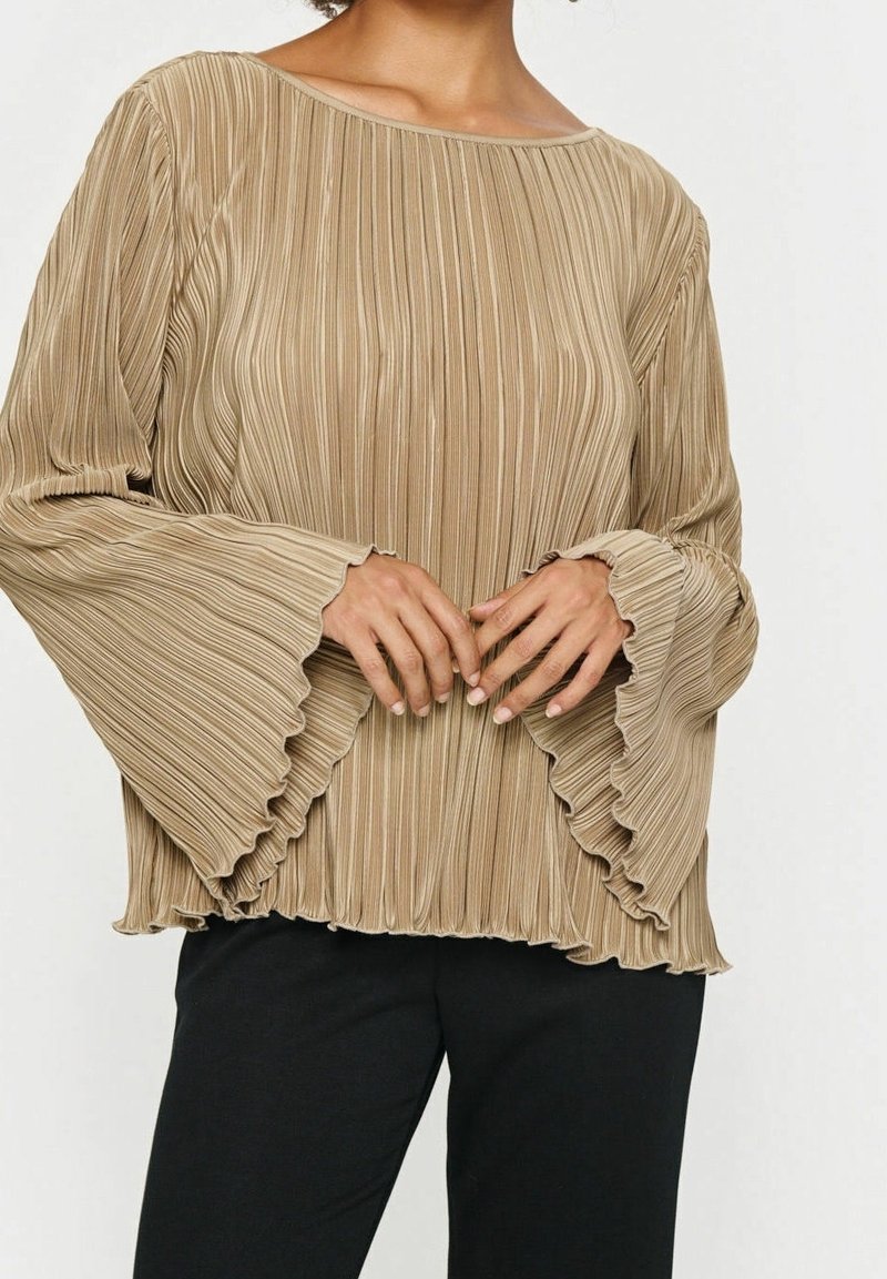 Femme portant un chemisier beige plissé à manches longues avec des poignets évasés volantés et un pantalon noir, les mains doucement jointes devant.