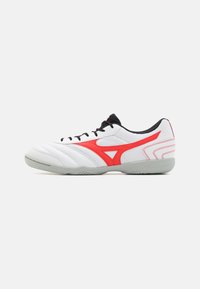 Scarpa sportiva bianca con un design laterale rosso, dettagli neri e una tomaia testurizzata. Presenta una suola in gomma grigia e lacci neri.