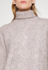 Jasnoszary sweter w splot warkoczowy z wysokim kołnierzem, wykonany z miękkiego, teksturowanego materiału z widocznym detalem wzoru na całej powierzchni.