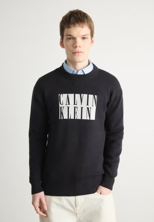 Pull en maille noir avec un col rond et des manches longues, arborant un grand logo blanc Calvin Klein sur la poitrine. Texture douce, coupe décontractée.