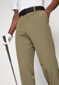 Muškarac u khaki hlačama i crnim remenom drži golf palicu s rukom u rukavicama, dok je druga ruka u džepu, na ravnoj pozadini.