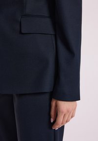 Zblizenie na granatową marynarkę skrojoną na miarę, z teksturowanym materiałem, wyraźnym kołnierzem i eleganckim krojem, na którym spoczywa dłoń.