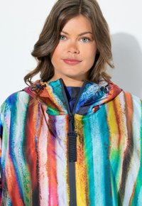 Jeune femme aux cheveux châtain clair portant une veste à capuche rayée multicolore avec une fermeture éclair noire dans un portrait en gros plan.