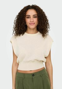 Cremfarbener, cropped Pullover aus weichem Strick mit kurzen, umgeschlagenen Ärmeln. Kombiniert mit grünen, plissierten Hosen mit einem Frontknopf.