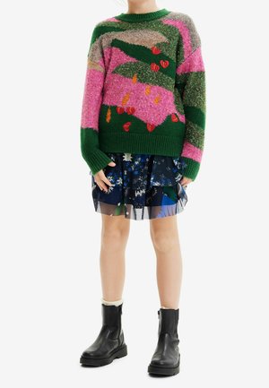 Enfant portant un pull à motifs verts et roses, une jupe fleurie à volants, des chaussettes crème et des bottines noires, debout sur un fond blanc.