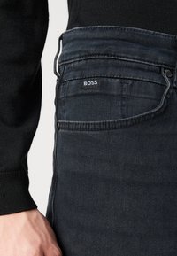 Tumedad denim püksid, millel on vormitud disain, väikese "BOSS" sildiga vööl ja siledat tekstuuri. Must kampsun on osaliselt nähtav.