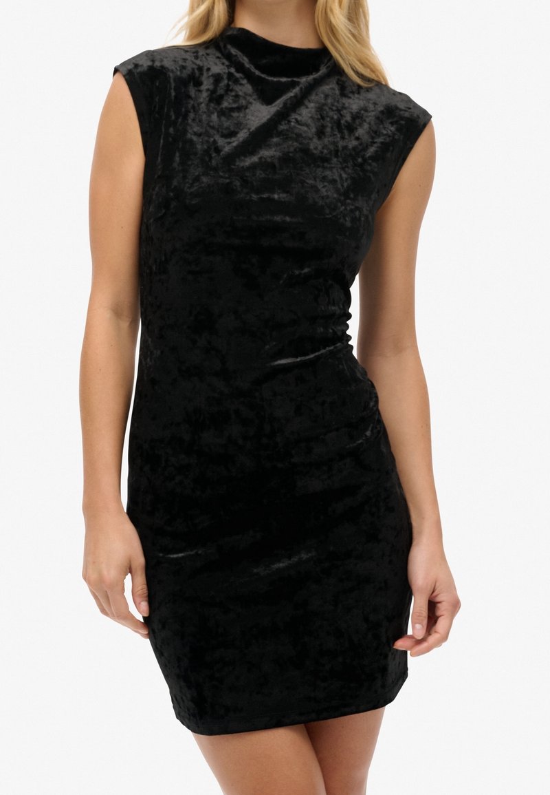 Femme portant une robe moulante en velours noir sans manches avec un col montant, debout devant un fond uni.