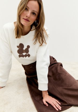 Witte sweatshirt met een bruine poedelafbeelding, gecombineerd met een bruine suède rok. Zacht materiaal en relaxte pasvorm, met een ronde hals.