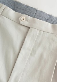 Gros plan de la ceinture d’un pantalon beige avec un seul bouton, une boucle de ceinture et une doublure intérieure à motif chevrons bleu et blanc.