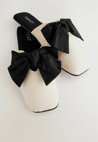 Pantofole beige senza lacci con una finitura testurizzata, caratterizzate da grandi fiocchi in tessuto nero sulla parte anteriore e suole nere. Logo del brand visibile.