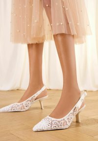 Beli čipkasti čevlji s peto z vrvico, z ostrimi konicami in dekorativnim izrezom, z ozko peto in pentljo na zadnji strani.