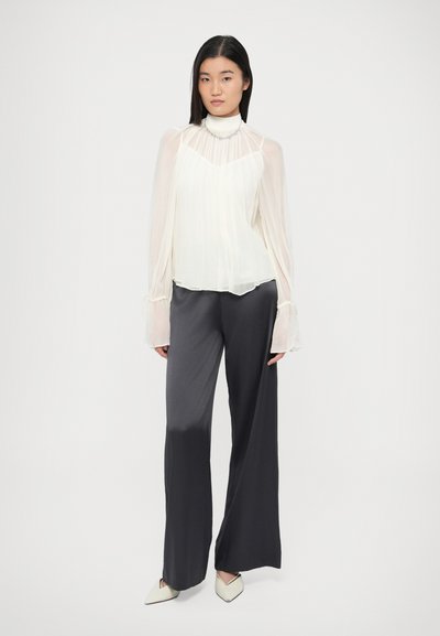 Blouse blanche transparente avec plis et manches évasées, associée à un pantalon large gris foncé et des escarpins blancs à bout pointu.