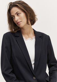 Navy blazer with structured shoulders and notched lapels, featuring a single button closure, worn over a white lace-trimmed top.