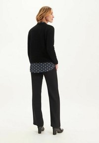Pull en maille noir avec une finition texturée, superposé sur une blouse bleue à motifs, associé à un pantalon noir et des bottes à talons.