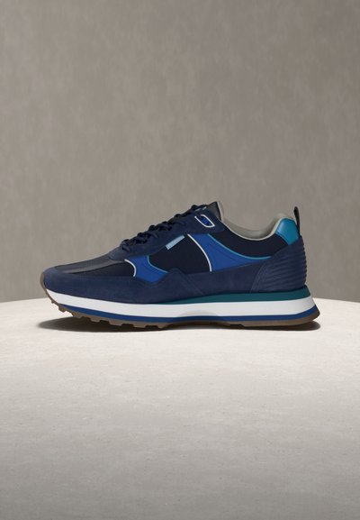 Tmavě modrý teniska s modrými a tyrkysovými akcenty, bílou stélkou, hnědou podrážkou a černými tkaničkami na světlém kulatém povrchu na šedém pozadí.