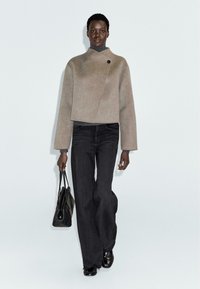 Beżowa, krótka wełniana kurtka z podwyższonym kołnierzem, z zapięciem na guziki, w połączeniu z czarnymi spodniami dzwonami i czarną torebką.