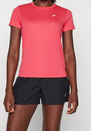 Femme portant un t-shirt de sport à manches courtes corail et un short de course noir, debout devant un fond uni.
