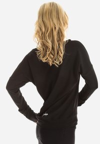 Čierny dlhý rukáv sweatshirt s voľným strihom, zaobleným spodným lemom, jemnou textúrou a logom na dolnej ľavej strane chrbta.