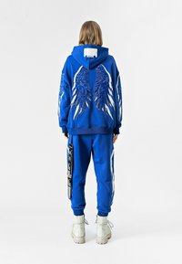 Sudadera azul con diseños de alas blancas y una etiqueta en la parte posterior, combinada con pantalones de chándal azules a juego que presentan detalles laterales y un tirante.