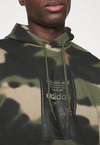 Grön och beige hoodie med kamouflagemönster, dragsko i huvan och en svart märkeslapp med präglad logotyp framtill. Tillverkad av mjukt tyg.