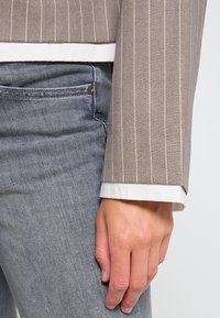 Bruine blazer met streepdessin en witte manchetten, gecombineerd met grijze spijkerbroek. Close-up van een hand die op de spijkerbroek rust, met zichtbare textuur en details.