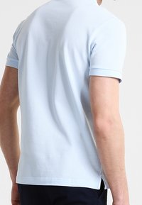 Man som bär en ljusblå kortärmad polo och mörka byxor, sedd från ryggen mot en enkel ljus bakgrund.