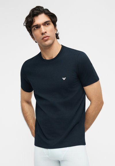 Άνδρας με σκούρα μαλλιά που φοράει στενή μαύρη κοντομάνικη μπλούζα με μικρό λευκό λογότυπο στο στήθος, στέκεται μπροστά σε απλό φωτεινό φόντο.