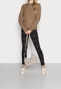 Frau trägt einen braunen Pullover, dunkelkarierte Hose, beige klobige Slipper und hält eine beigefarbene Handtasche mit einer Kettenhenkel vor ihr.