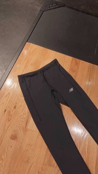 Sorte treningsbukser laget av glatt stoff med sidelommer og elastisk midjebånd, med en liten hvit logo på venstre lår.