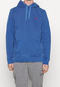 Sudadera de algodón azul con bolsillo canguro frontal, capucha con cordón y logo rojo en el pecho. Presenta puños y dobladillo acanalados.