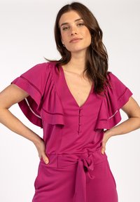 Fuchsia jurk met fladder mouwen, diepe V-hals en drie knoopdetails aan de voorkant. Inclusief een tailleband en een zachte textuur.