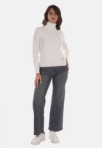 Maglione bianco a collo alto con una texture liscia e dettagli luccicanti, abbinato a jeans in denim scuri a vita alta e sneakers bianche.