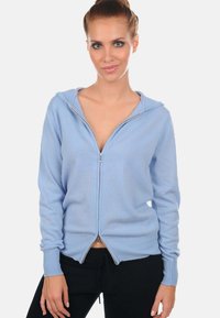 Haut à fermeture éclair bleu fabriqué en tissu doux, doté d'une capuche, de poignets côtelés et d'un design à ourlet fendu. Texture lisse sans motifs visibles.