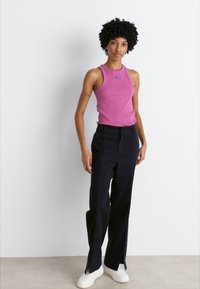 Top rosa senza maniche con logo, abbinato a pantaloni larghi scuri con spacchi laterali e sneaker bianche su uno sfondo neutro.