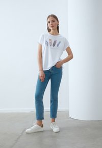 White cotton t-shirt featuring feather prints in various colors, paired with light blue jeans and white sneakers. Simple, relaxed fit.